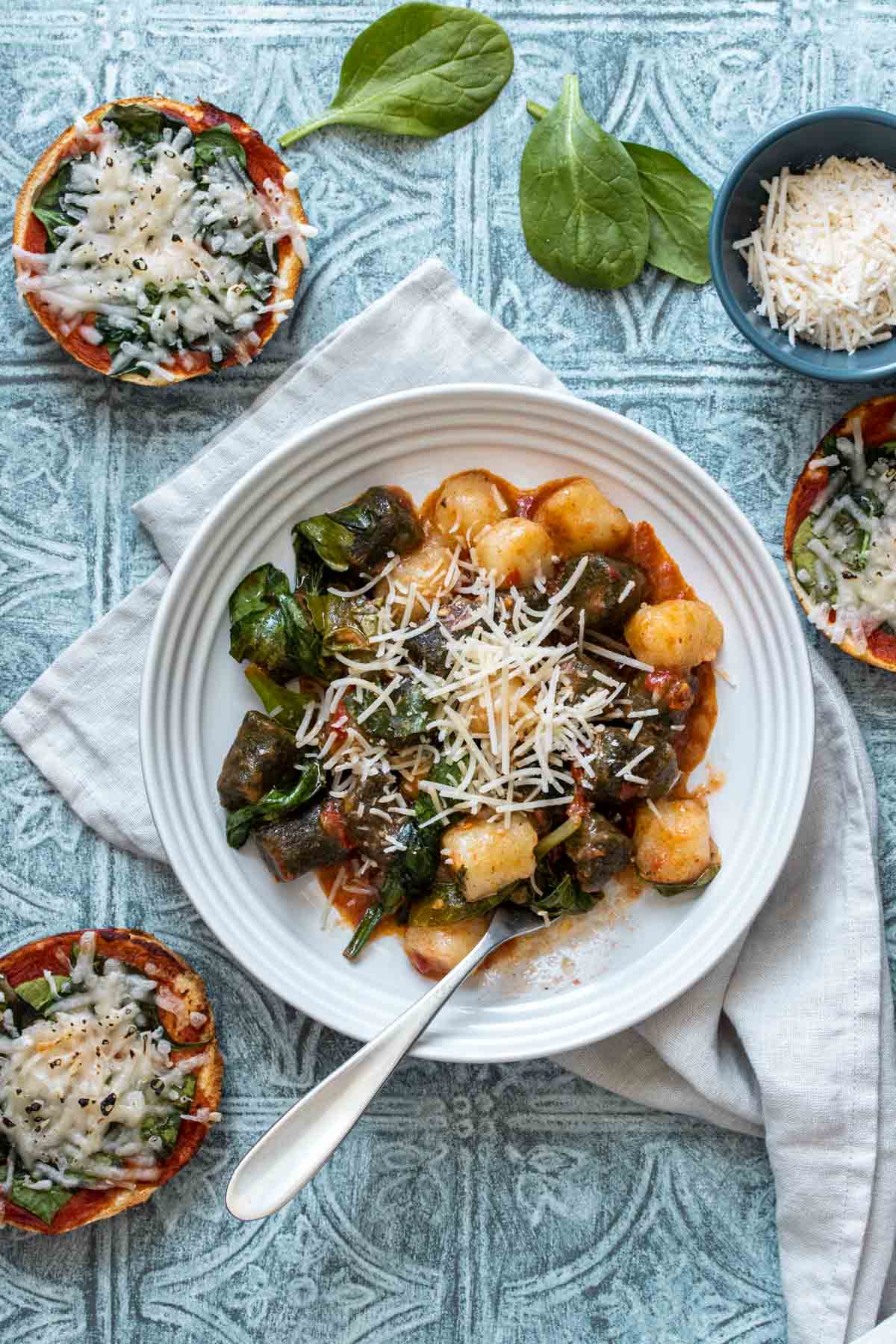 A white bowl with gnocchi, red sauce, spinach and parmesan on a blue surface with pizza bagels around it.