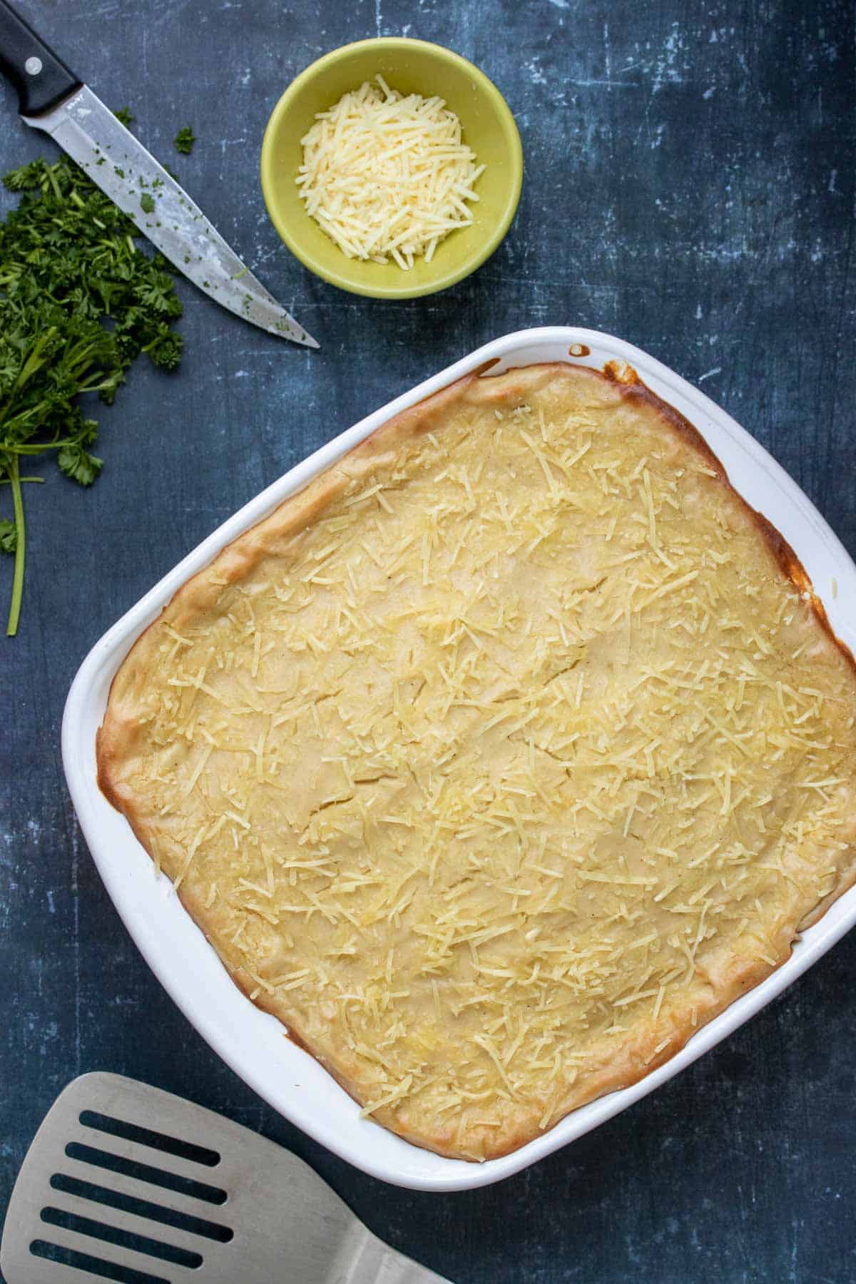 Top view of a baked casserole like dish with a creamy top covered in Parmesan next to a metal spatula