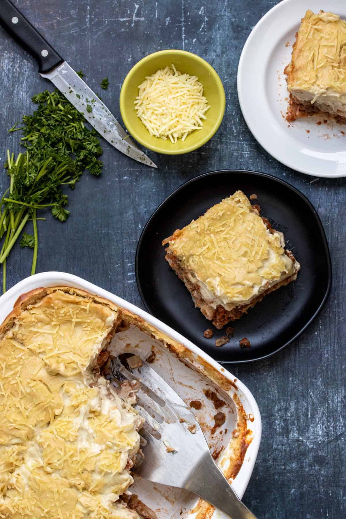 Top view of a baking dish with pastitsio and a silver spatula next to plates of pieces of it