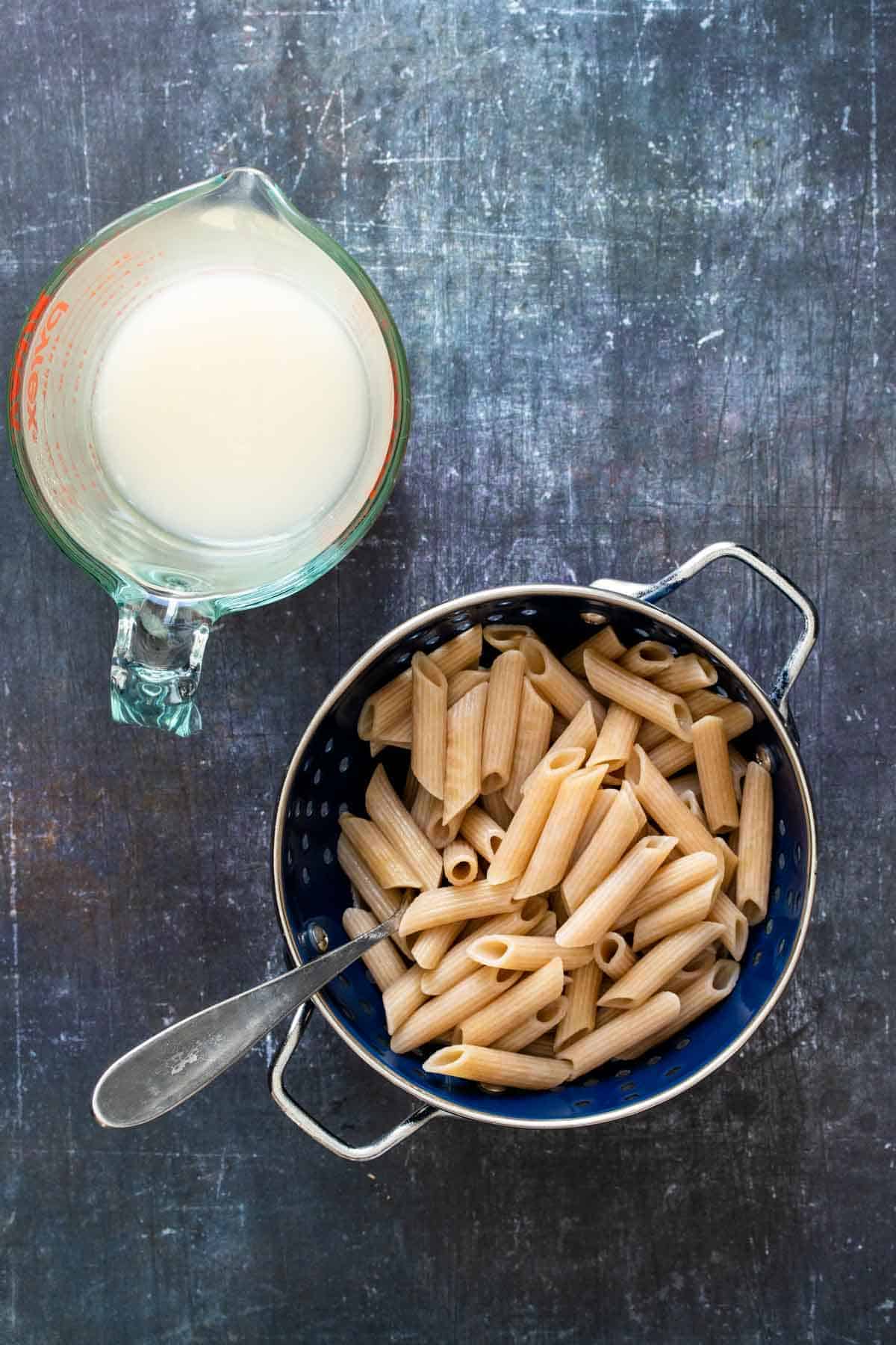 Top view of cooked pasta in a strainer next to a glass cup with pasta water