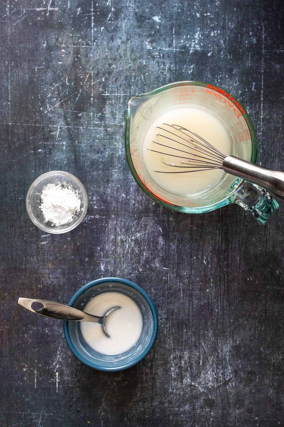 A blue bowl with a milky liquid next to tapioca starch in a small bowl and pasta water in a glass cup