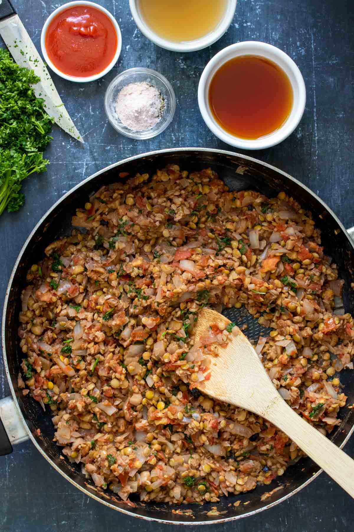 Wooden spoon mixing a chopped bean and tomato mixture in a large pan next to bowls of ingredients