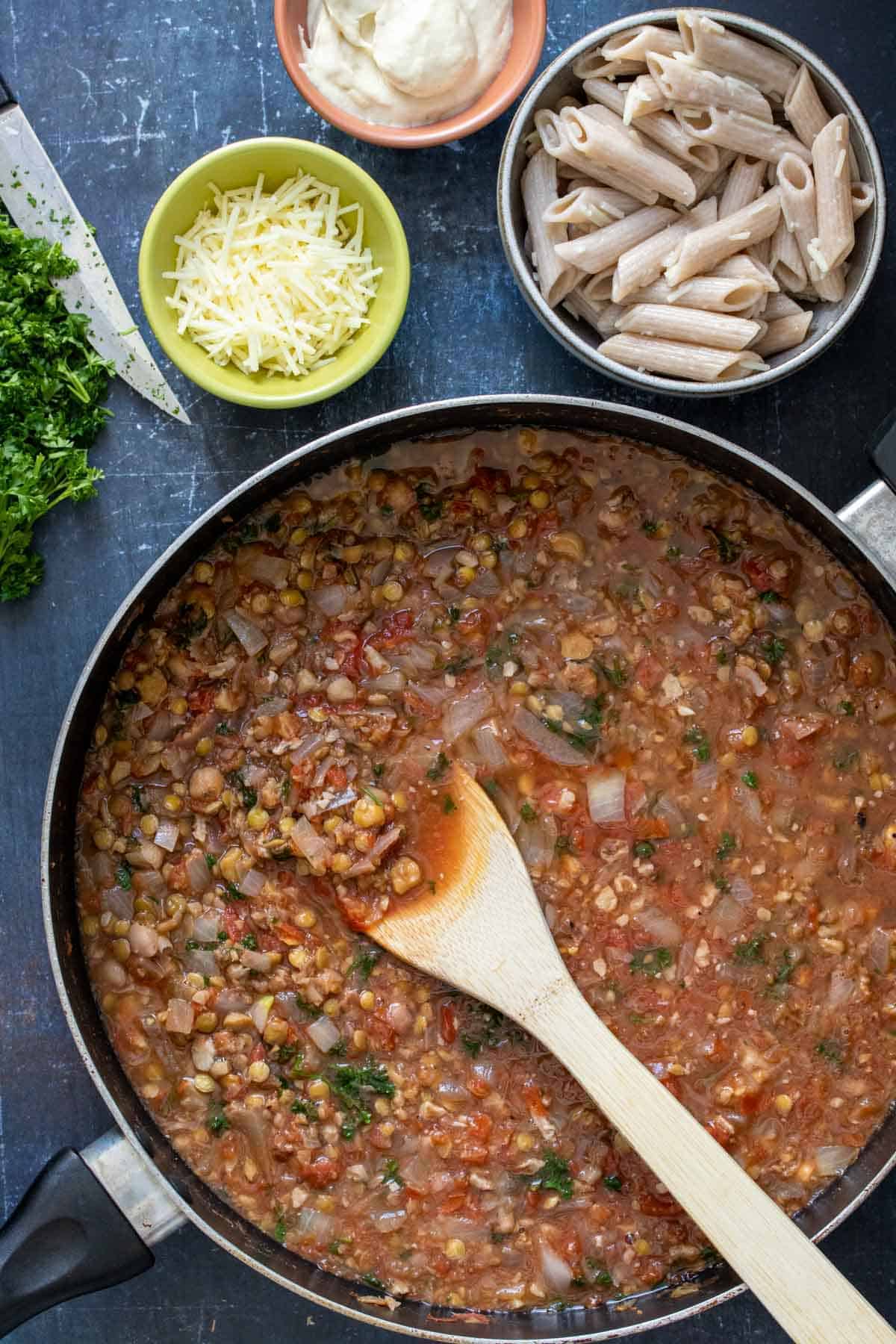 A wooden spoon mixing a liquidy mixture of beans and tomatoes next to bowls of pasta, cheese and bechamel