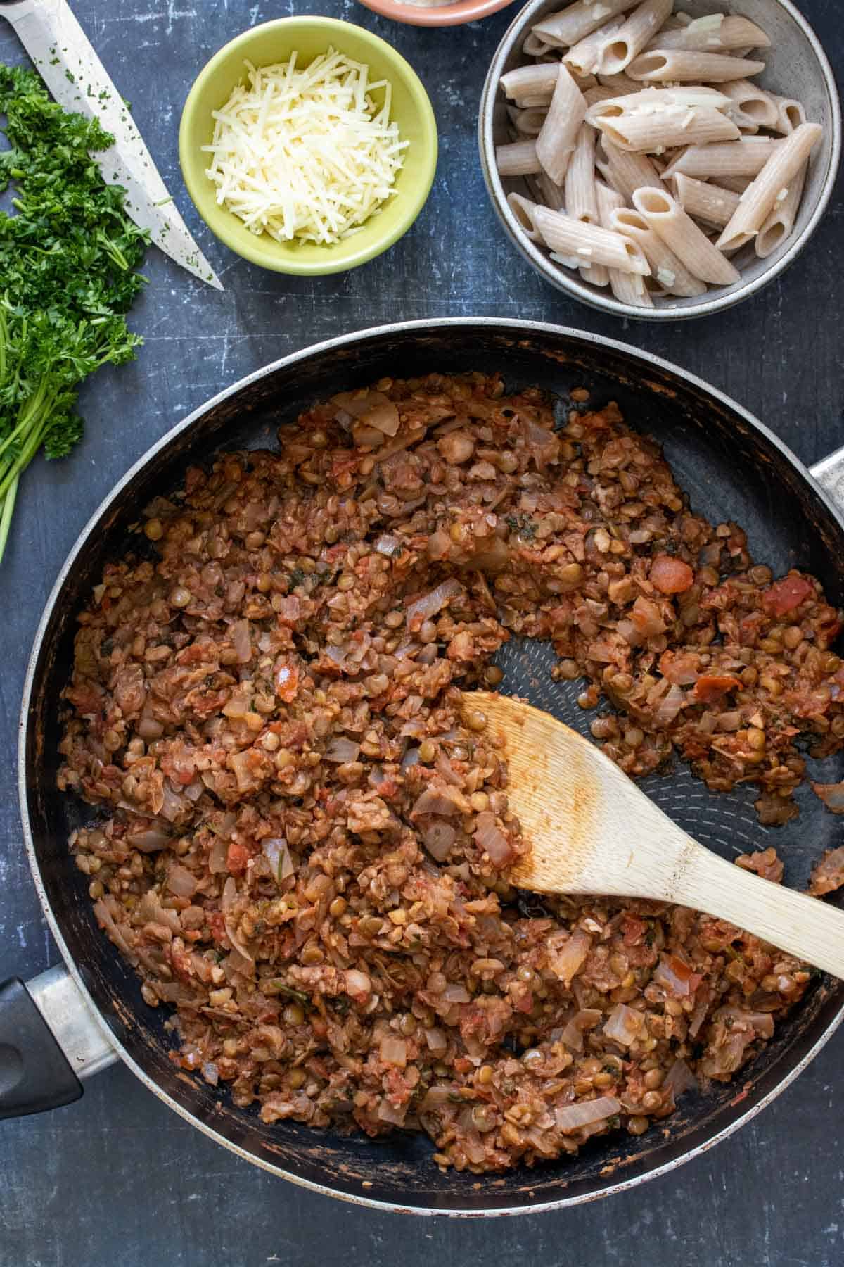 Top view of a meaty looking tomato and bean mixture being stirred by a wooden spoon next to bowls of ingredients