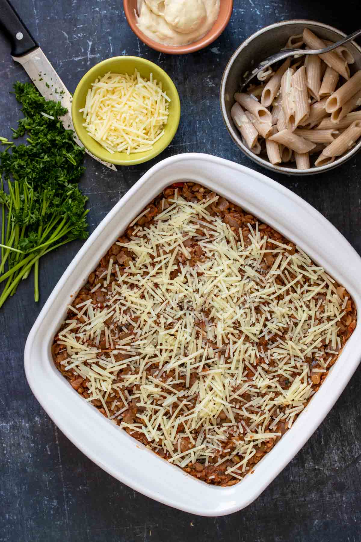 A white baking dish with Parmesan spread over a meaty looking tomato based mixture next to bowls of pasta, cheese and Bechamel