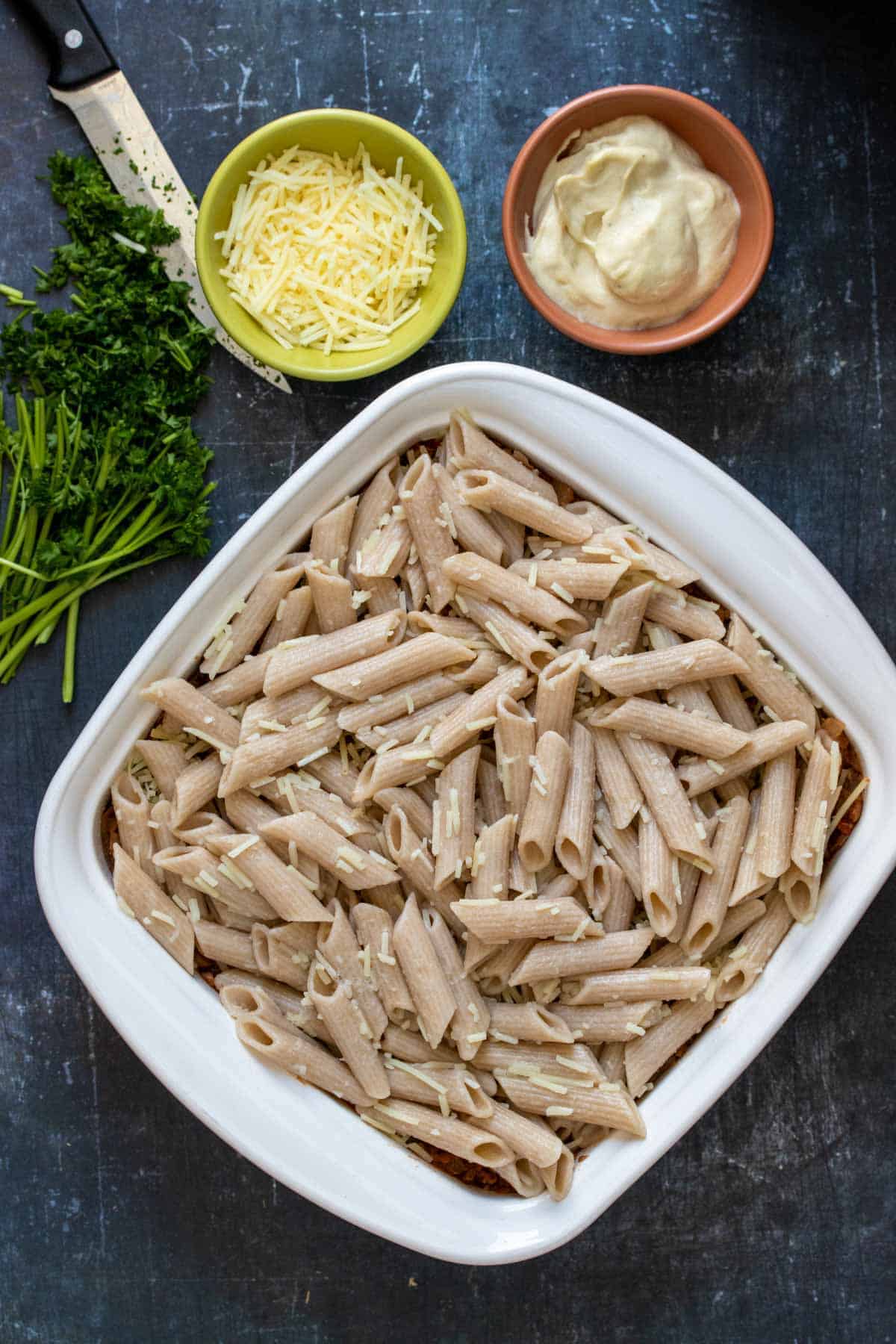 Top view of a white baking dish with pasta filled inside next to chopped parsley and bowls of Parmesan and Bechamel