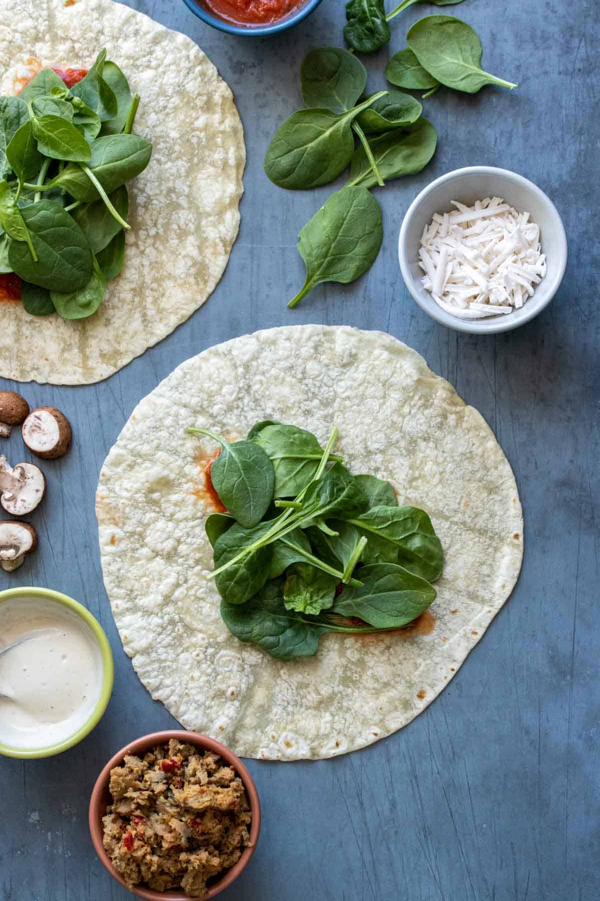 A slate surface with a tortilla with spinach on it and other pizza ingredients around it