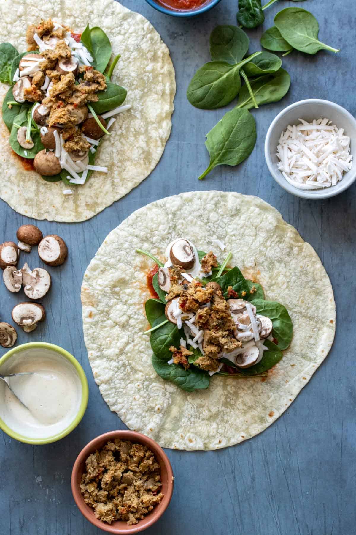 A tortilla on a slate surface with spinach, mushrooms, cheese and sausage and the ingredients around it too