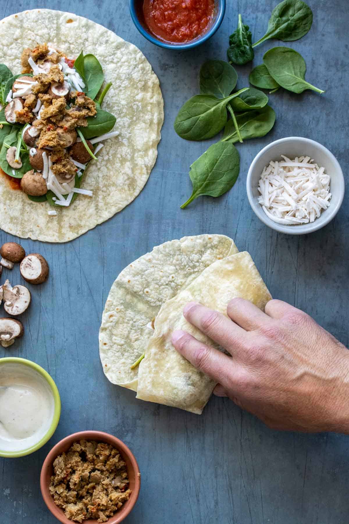 A hand rolling a burrito up next to a tortilla with pizza ingredients on top