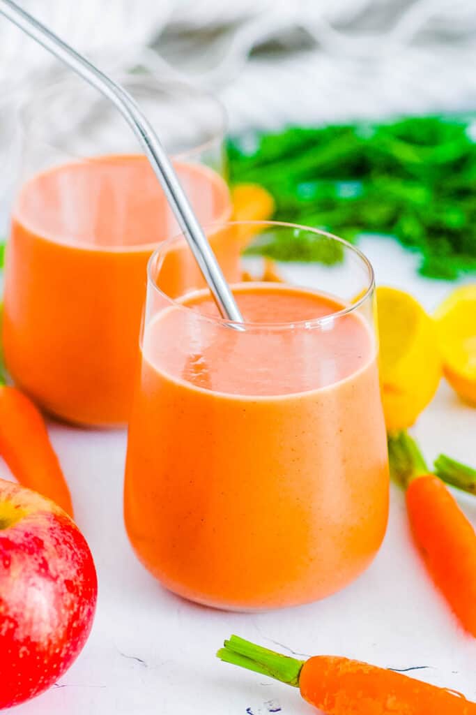 Two glasses of apple carrot smoothie on the counter next to napkins and fruit.