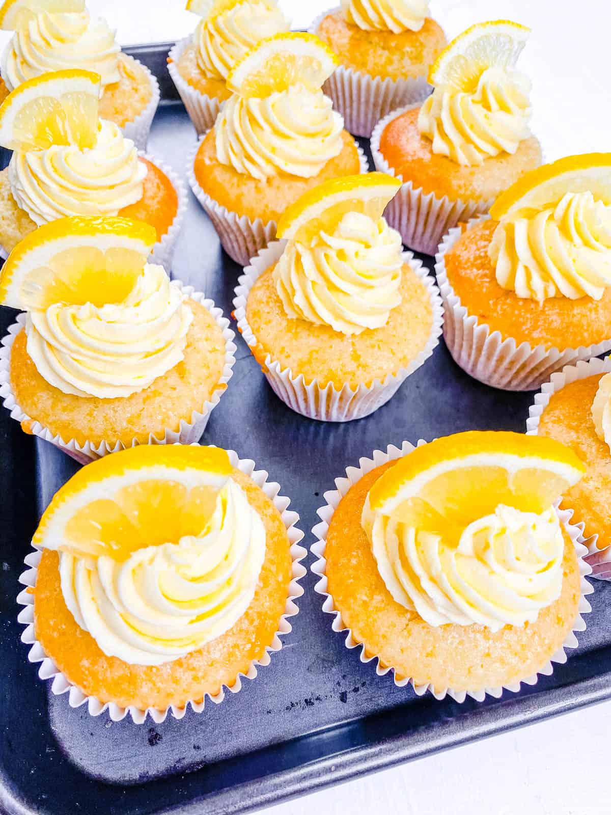 Vegan lemon cupcakes topped with vegan buttercream frosting and a lemon wedge on a baking sheet.