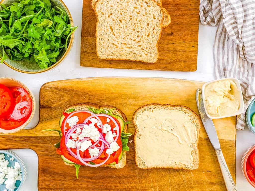 Whole wheat bread spread with hummus, topped with tomatoes, feta, onions, and greens on a cutting board.