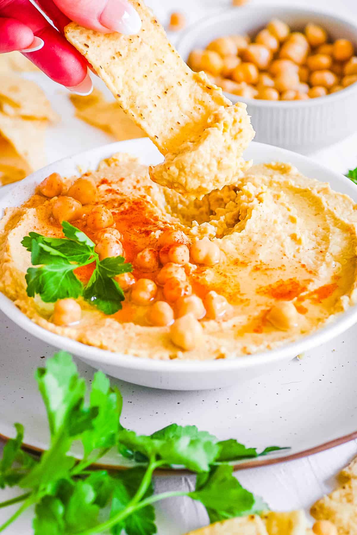 Hummus without garlic in a white bowl garnished with parsley and paprika, with a pita chip dipped in it.