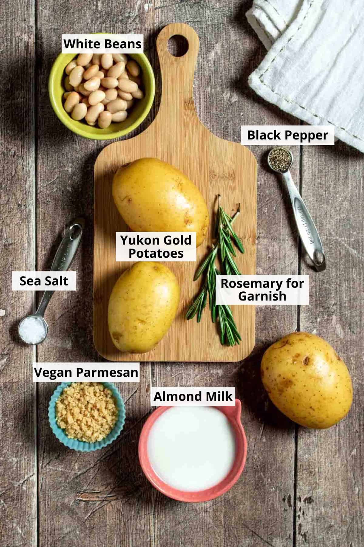 Top view of a wooden surface with potatoes and rosemary on a cutting board surrounded by beans, milk, parmesan and spices.