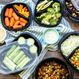 Fresh and cooked veggies in black containers of different sizes sitting on a striped towel with dips next to them.