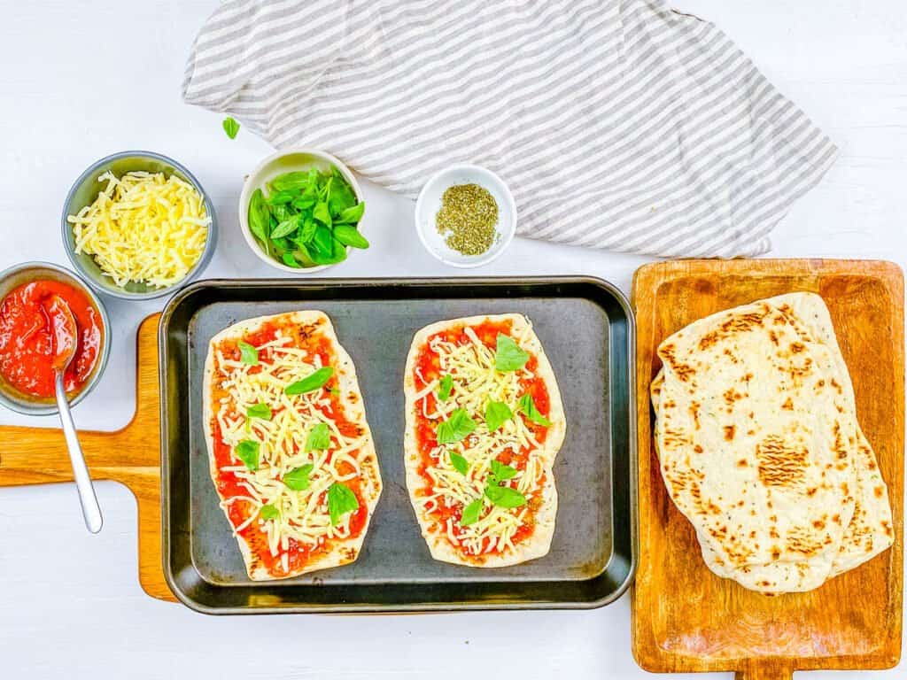 Naan topped with sauce, basil, and cheese on a wooden cutting board.