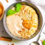Banana smoothie bowl topped with nut butter, banana slices, nuts and herbs in a grey bowl with a spoon.