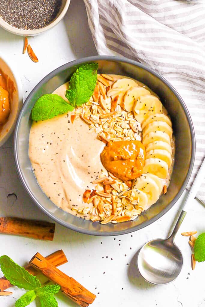 Banana smoothie bowl topped with nut butter, banana slices, nuts and herbs in a grey bowl with a spoon.