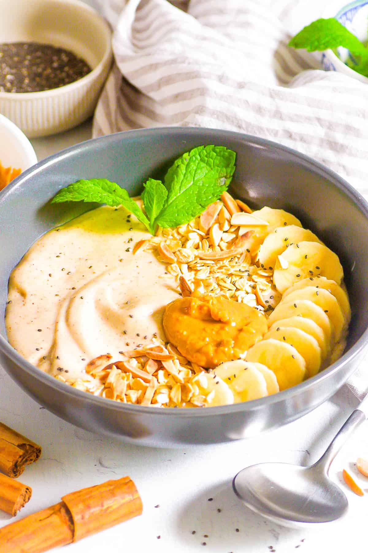 Banana smoothie bowl topped with nut butter, banana slices, nuts and herbs in a grey bowl with a spoon.