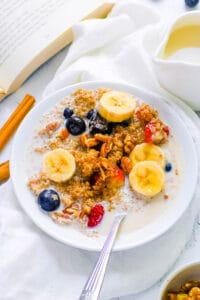 Quinoa oatmeal served with banana slices, nuts, milk and berries in a white bowl with a spoon.