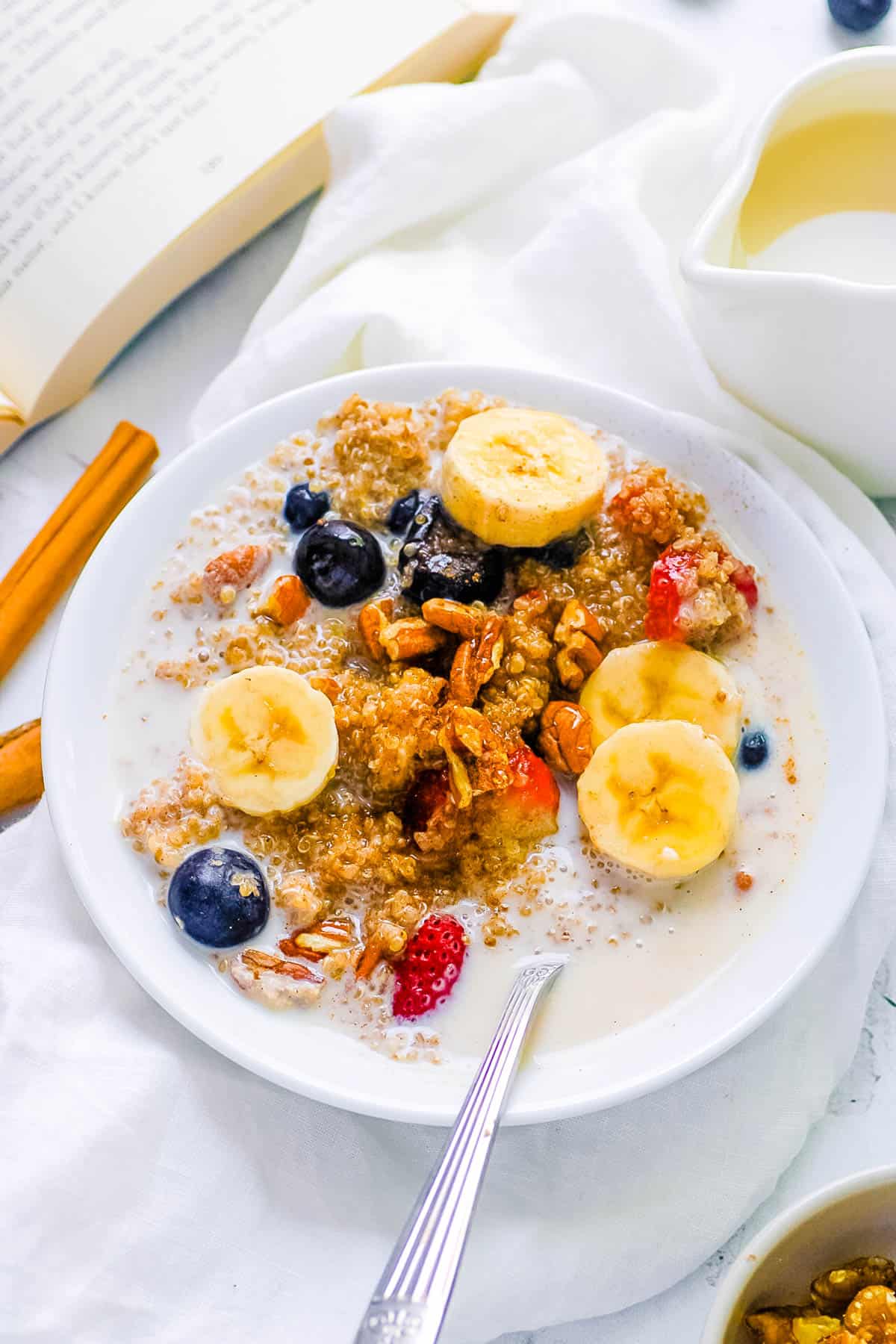 Quinoa oatmeal served with banana slices, nuts, milk and berries in a white bowl with a s،.