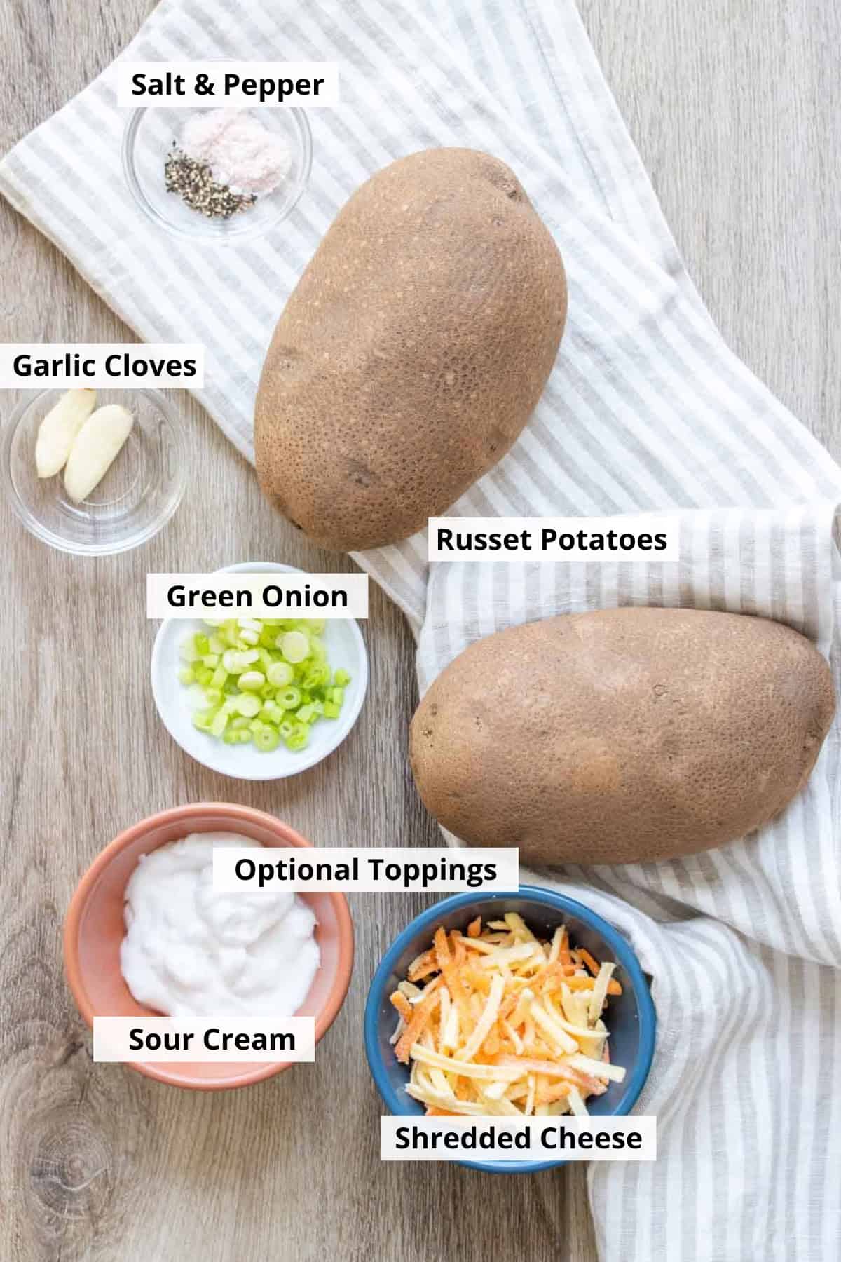 Whole potatoes, with bowls of cheese, sour cream, green onions, garlic, salt and pepper on a wooden surface