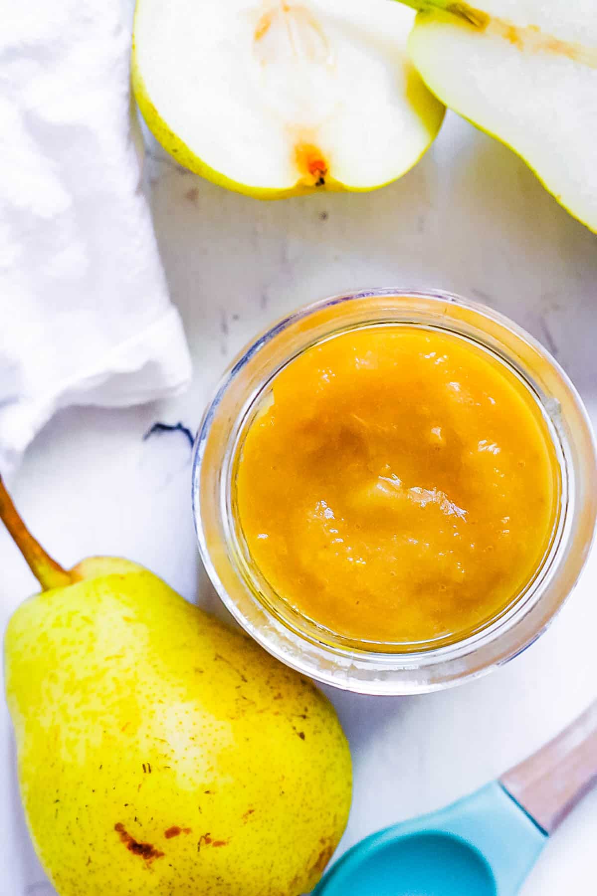 Pear baby food in a gl، jar on a white countertop with cut pears on the side.