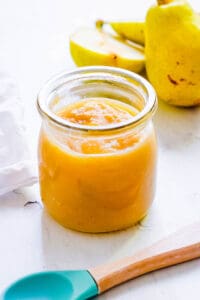 Pear puree baby food in a glass jar on a white countertop with cut pears on the side.