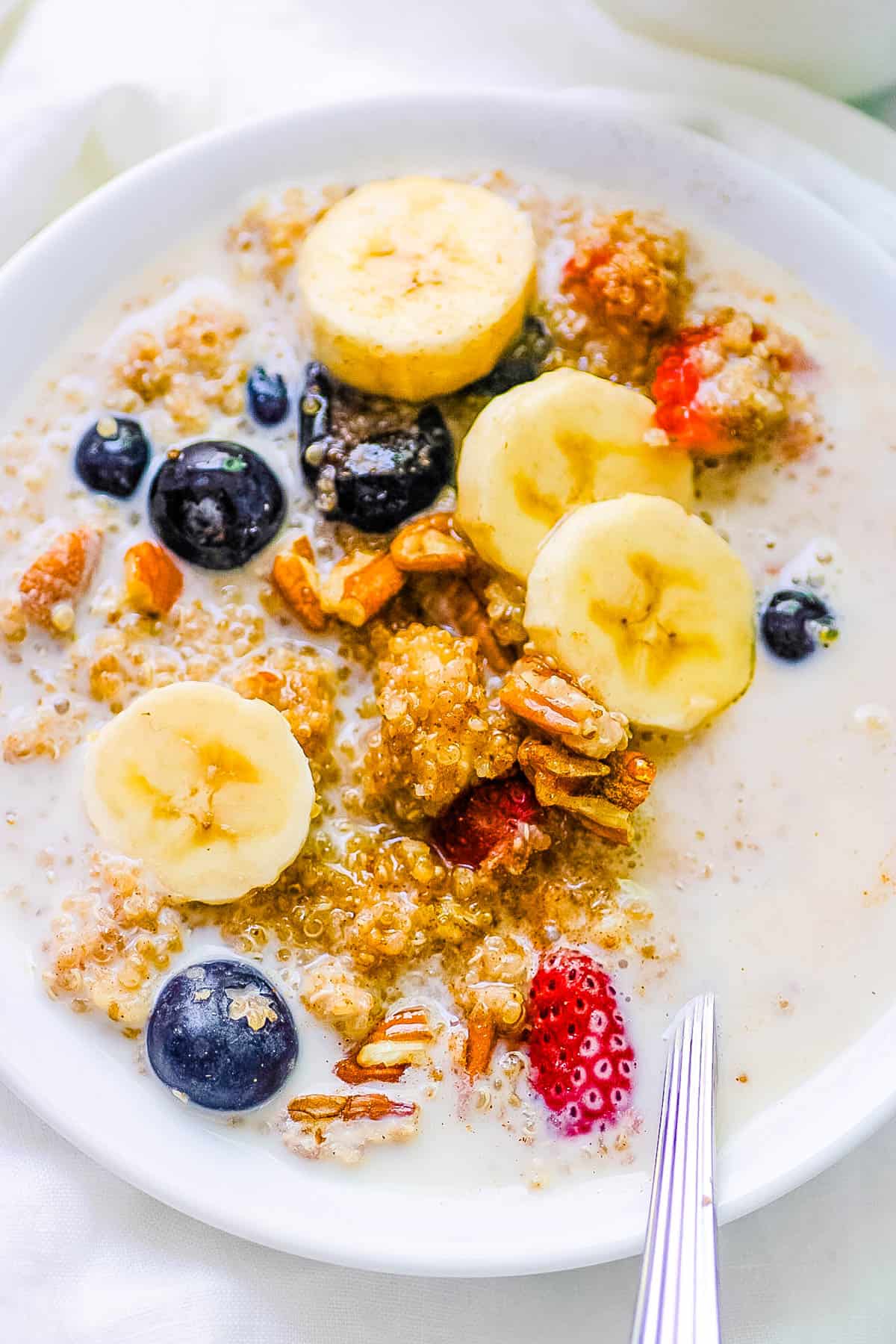 Quinoa oatmeal served with banana slices, nuts, milk and berries in a white bowl with a s،.