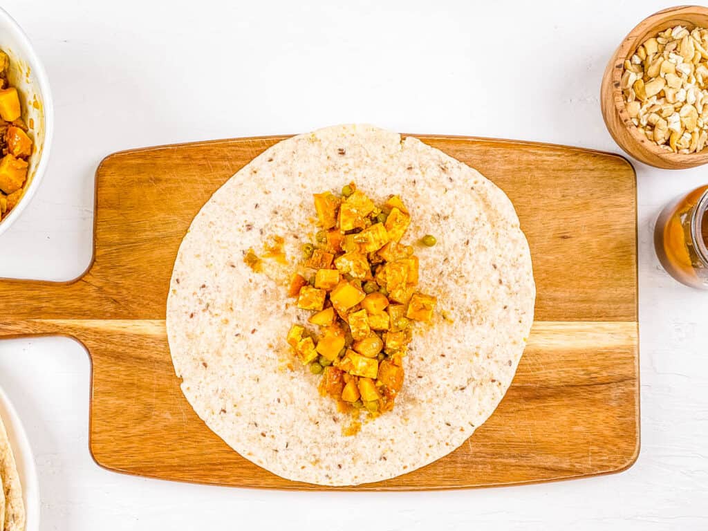 Potato tofu filling added to whole wheat tortilla on a wooden cutting board.