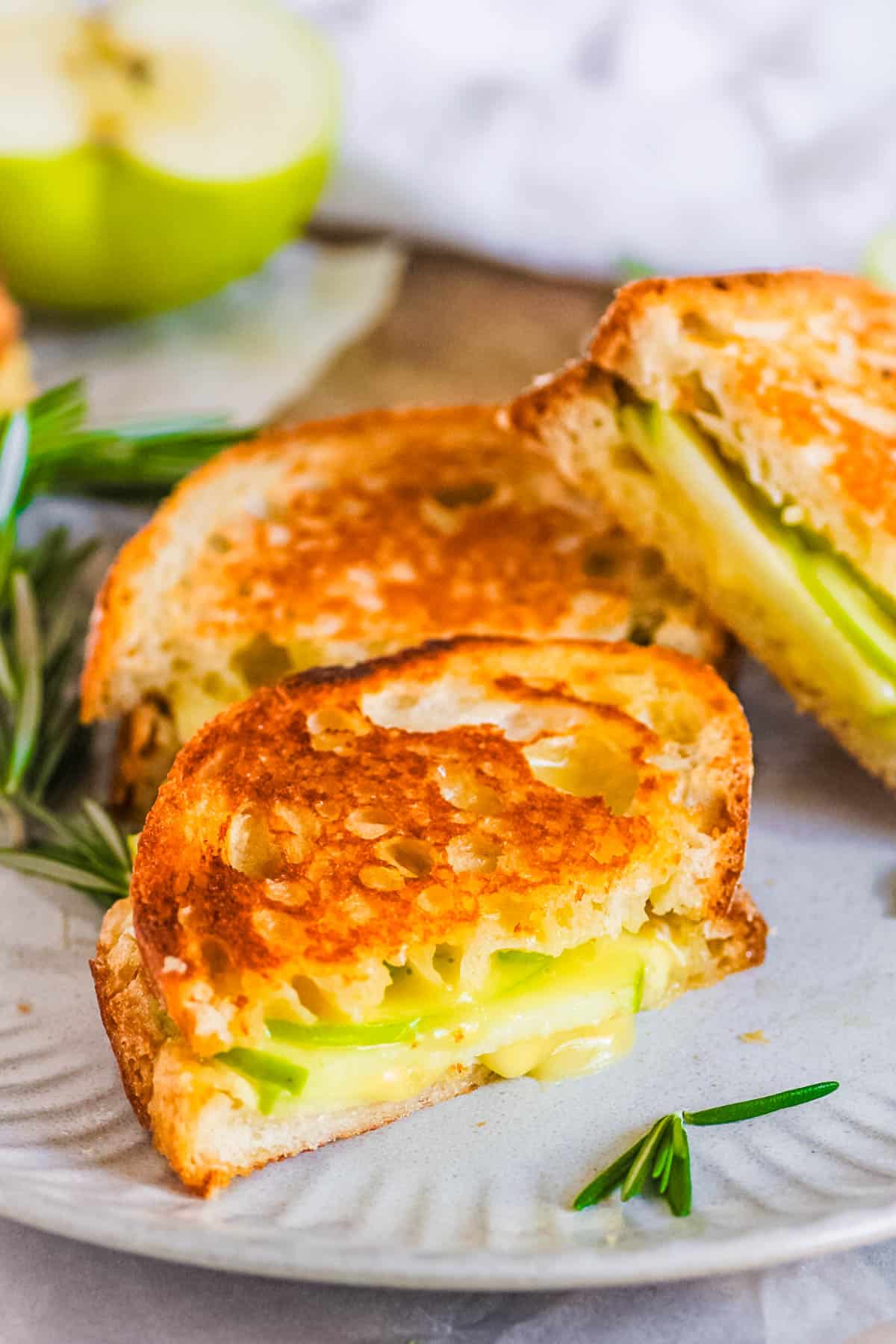 Sourdough grilled cheese sandwich with apple and brie cut and stacked on a plate with fresh herbs.