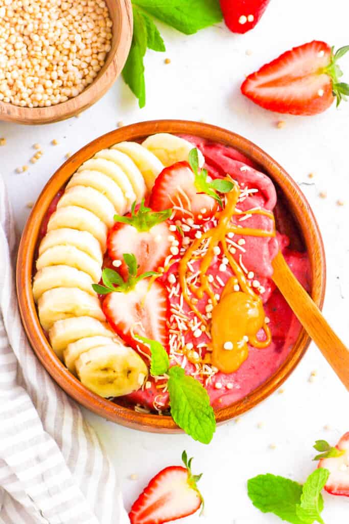 Strawberry smoothie bowl topped with banana slices, strawberries, nut butters and seeds on a white countertop.