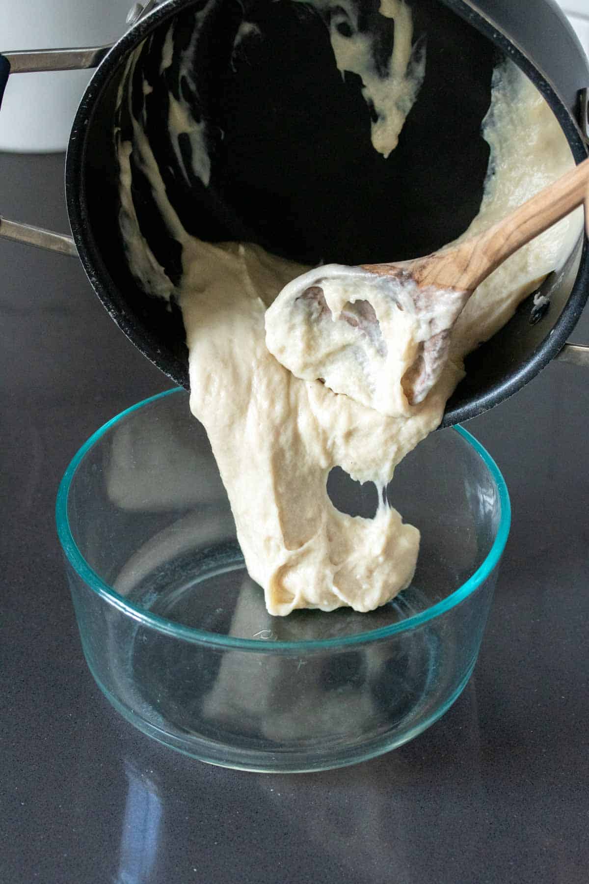 A wooden spoon scooping a thick cheese like substance from a black pot into a glass bowl.