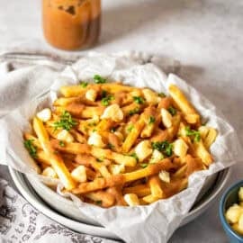 A stack of white bowls with parchment paper and vegan poutine style french fries in them next to a towel and jar of gravy.