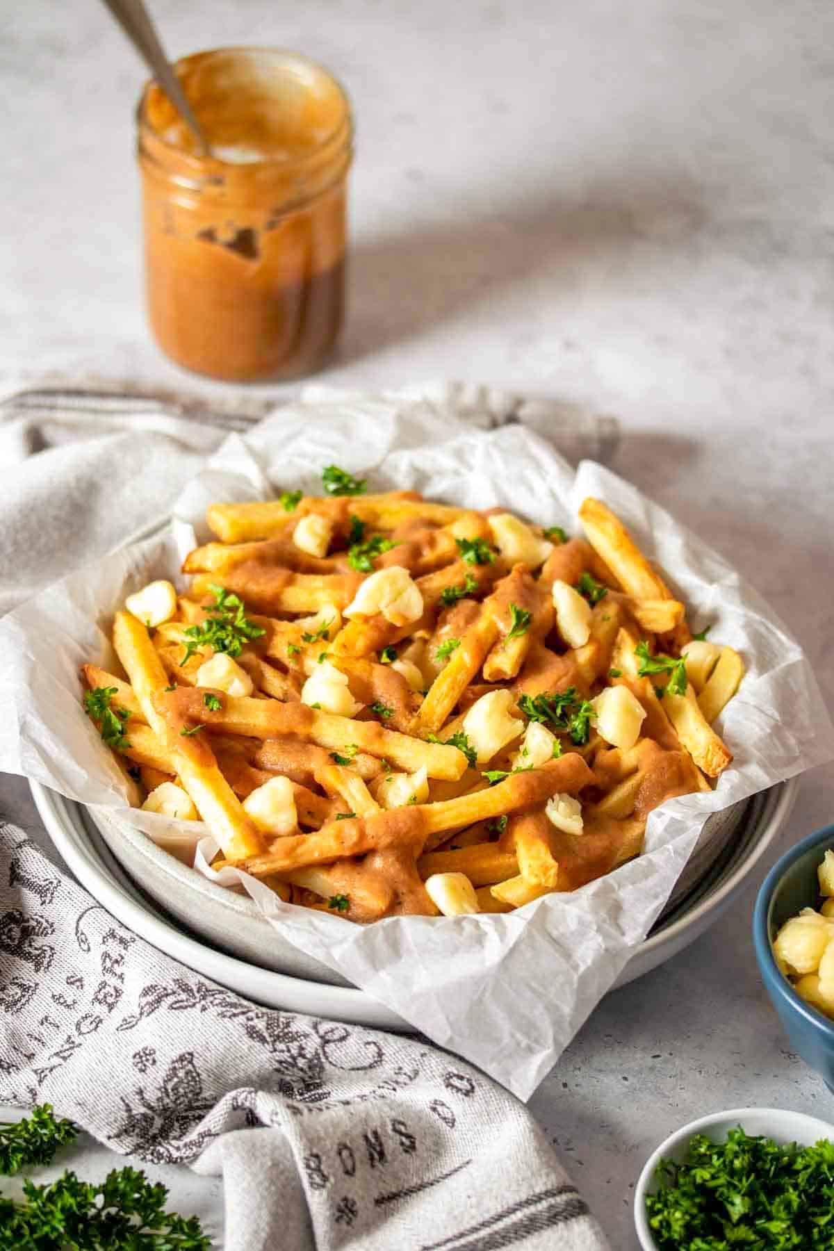 A stack of white bowls with parchment paper and vegan poutine style french fries in them next to a towel and jar of gravy.