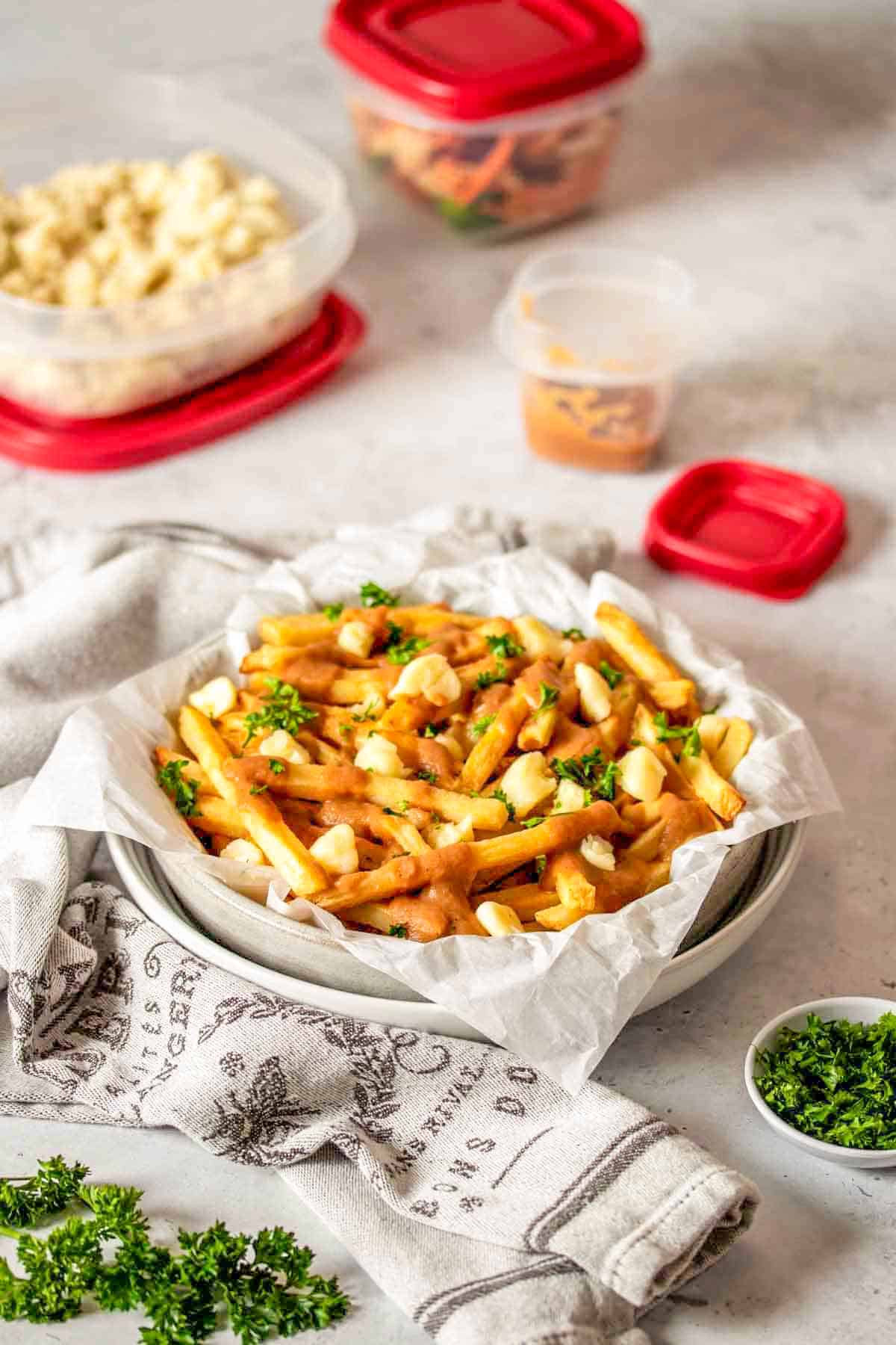 A stack of white bowls with parchment paper and vegan poutine style french fries in them next to a towel and jar of gravy.