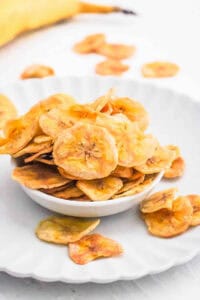 Air fryer banana chips in a white bowl, overflowing onto a white plate.