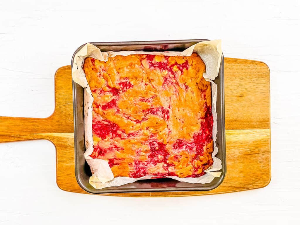 Baked raspberry blondies in a baking tin on a cutting board.