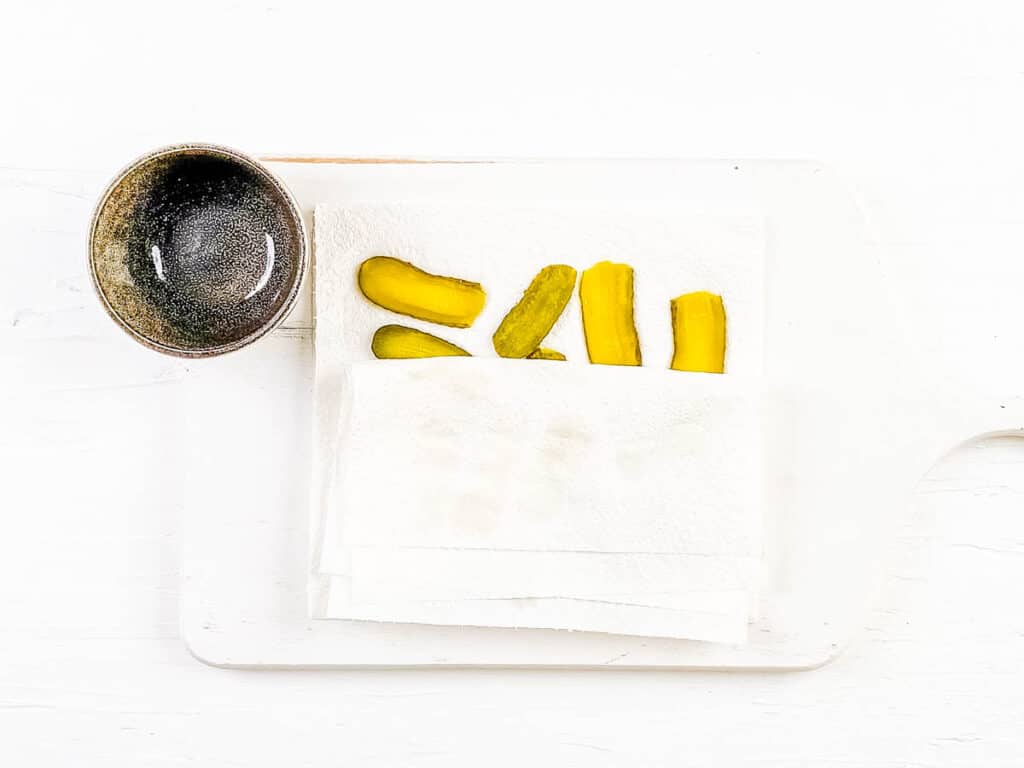 Dill pickles being blotted between paper towels on a cutting board.