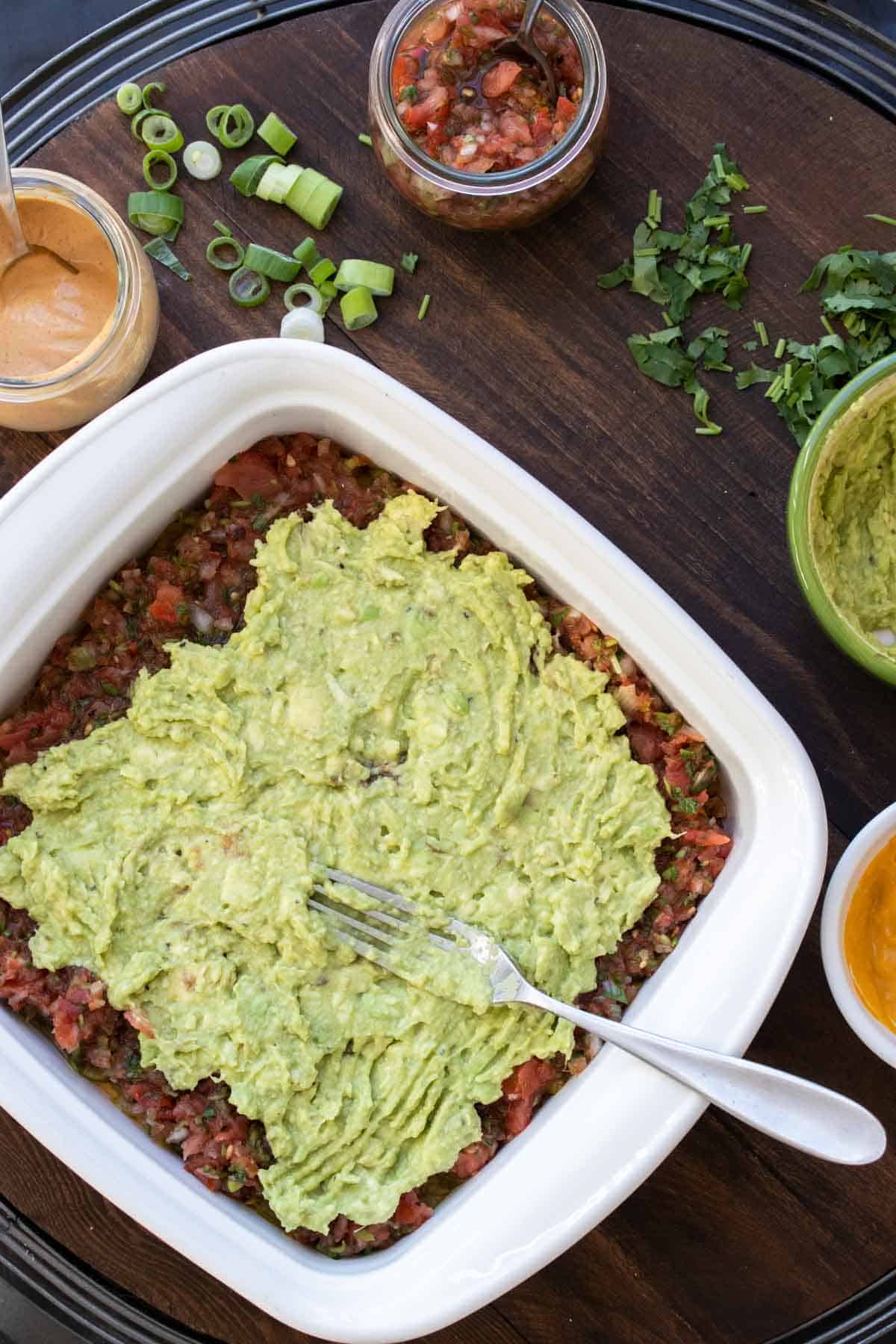 A fork spreading guacamole over a layer of salsa that is topped on a 7 layer dip in a white baking dish.