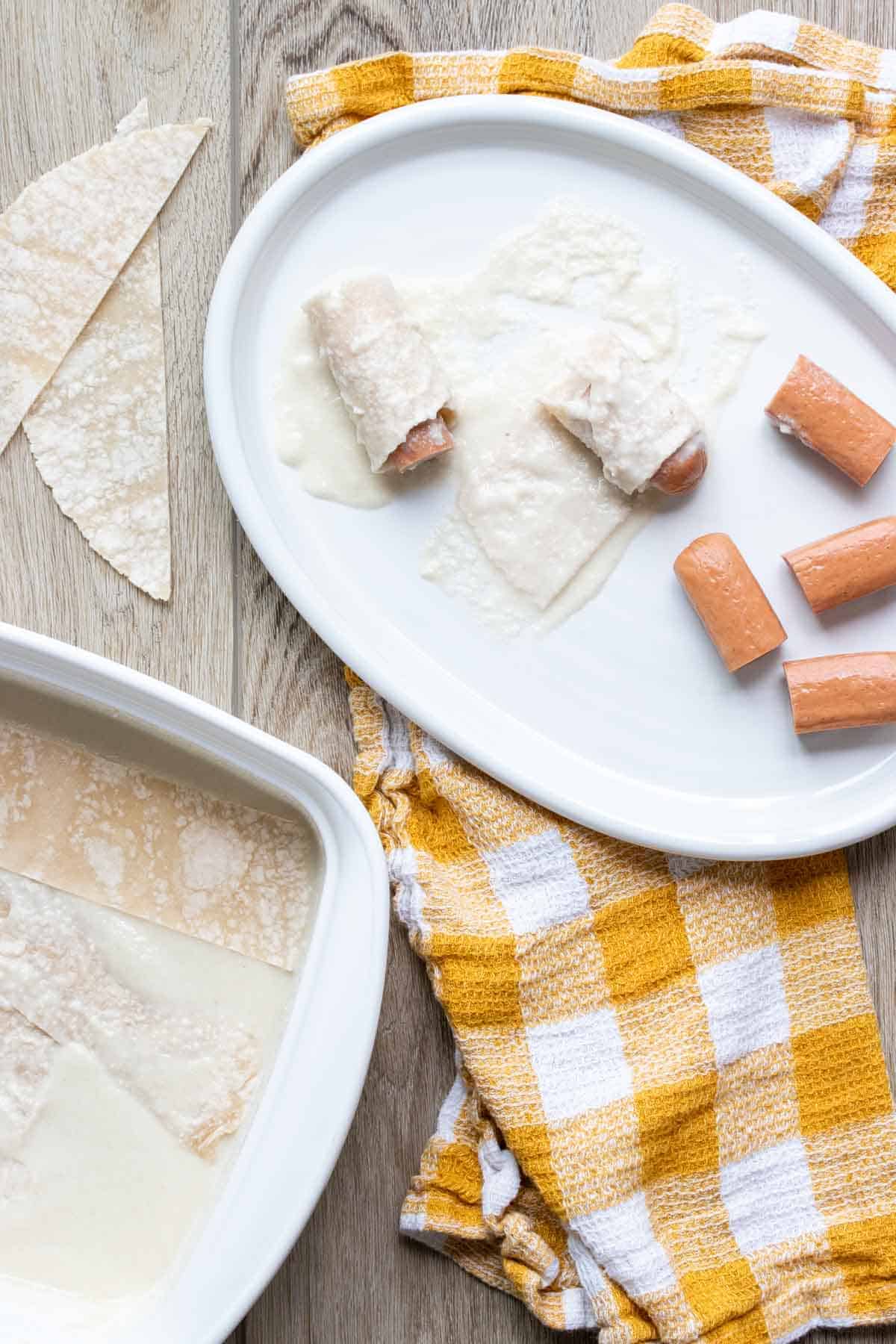 A white platter with pieces of hot dog and some rolled with batter coated tortilla strips.