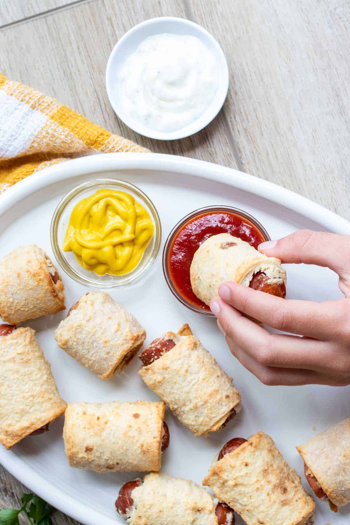 Hand dipping a pig in a blanket into a dipper of ketchup.