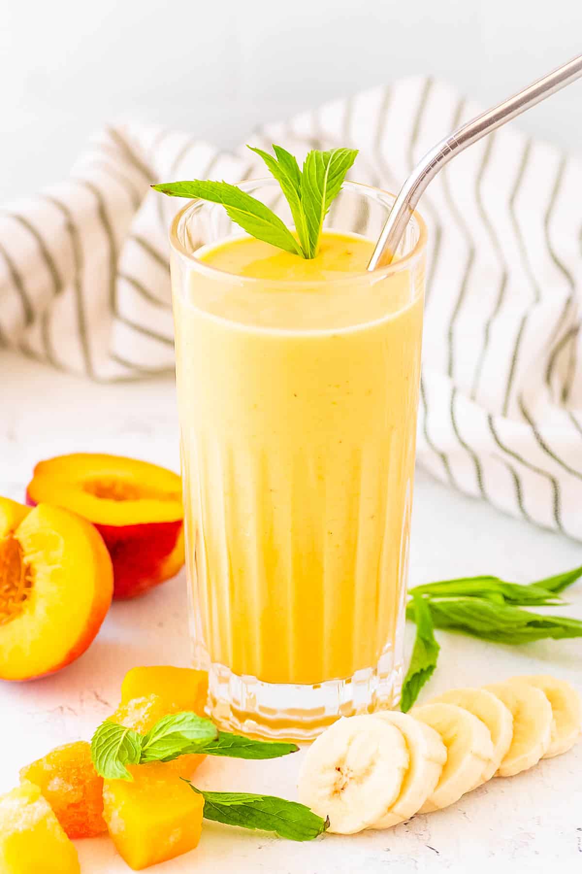 Peach banana smoothie served in a gl، with a straw and mint leaves as a garnish.