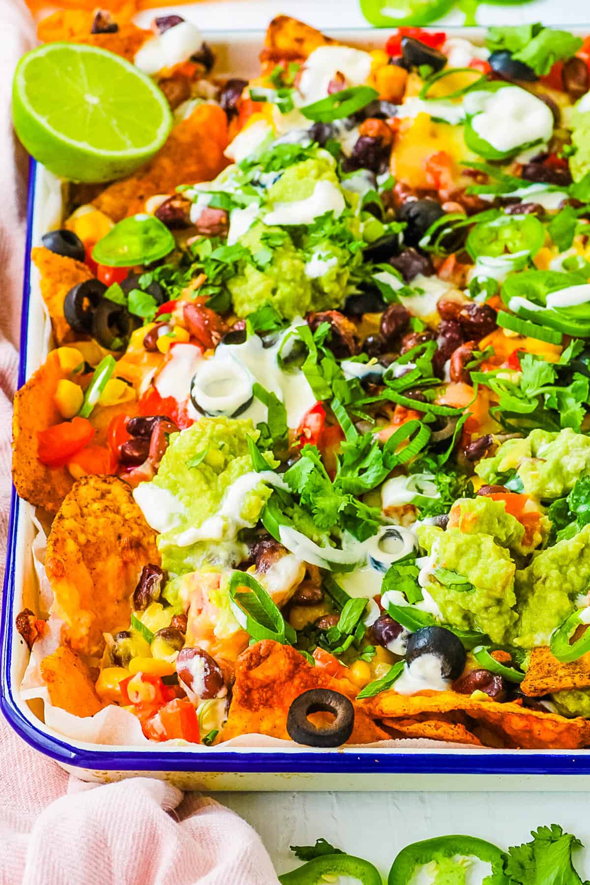 Loaded vegetarian nachos piled with veggies, jalapenos, guacamole, sour cream, cheese, olives served on a white platter.