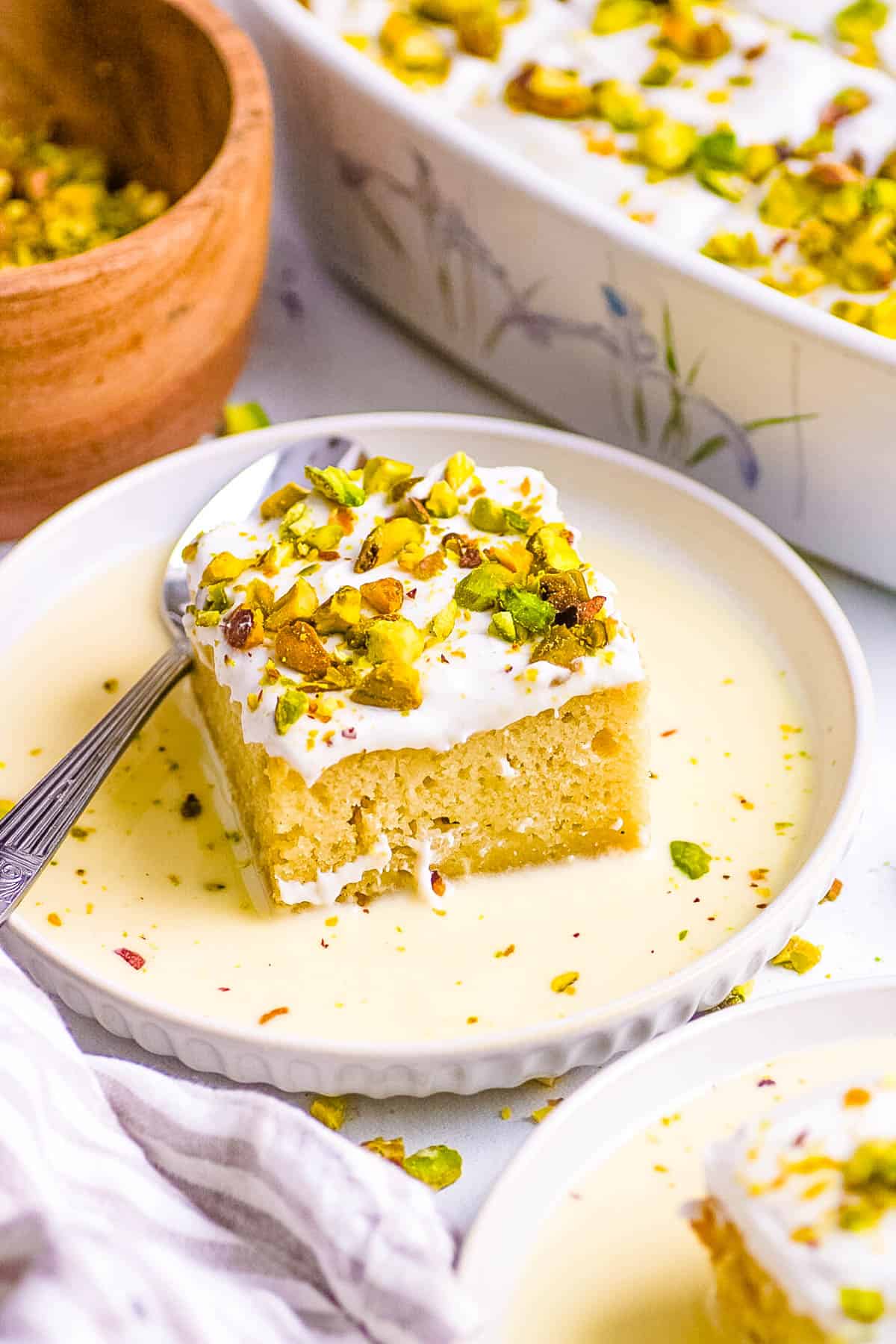 Rasmalai tres leches cake served on a white plate, topped with pistachios and the sweetened milk mixture.