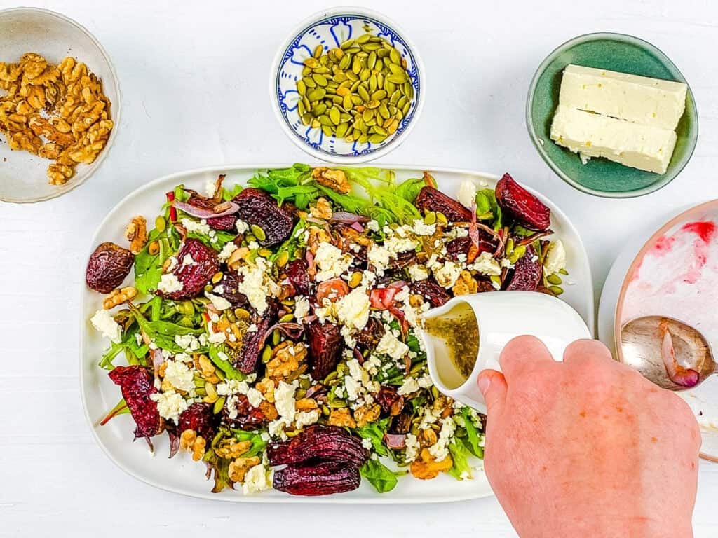 Salad dressing added to roasted beetroot and feta salad with walnuts on a white platter.