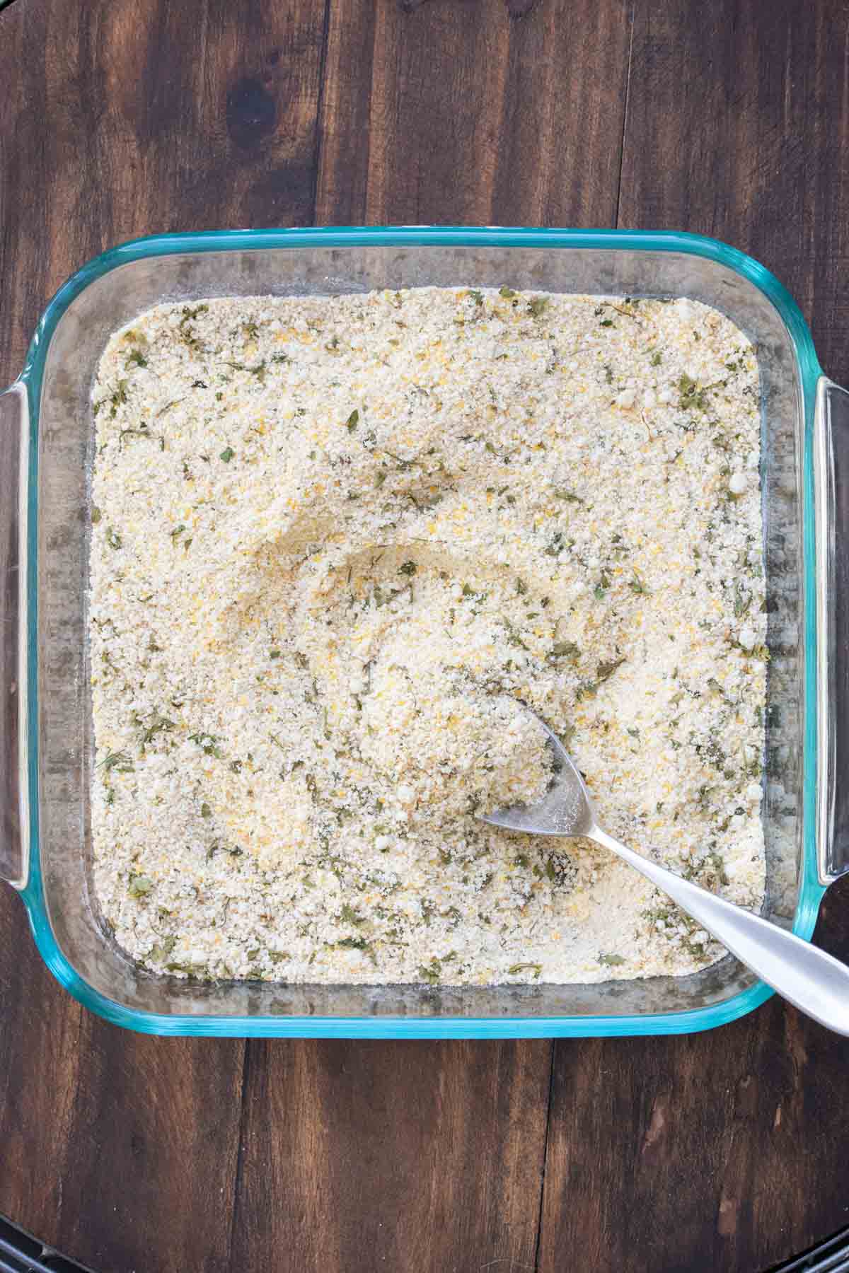 A square glass baking dish with a mixture of flour, cornmeal and spices in it and a spoon picking some up.