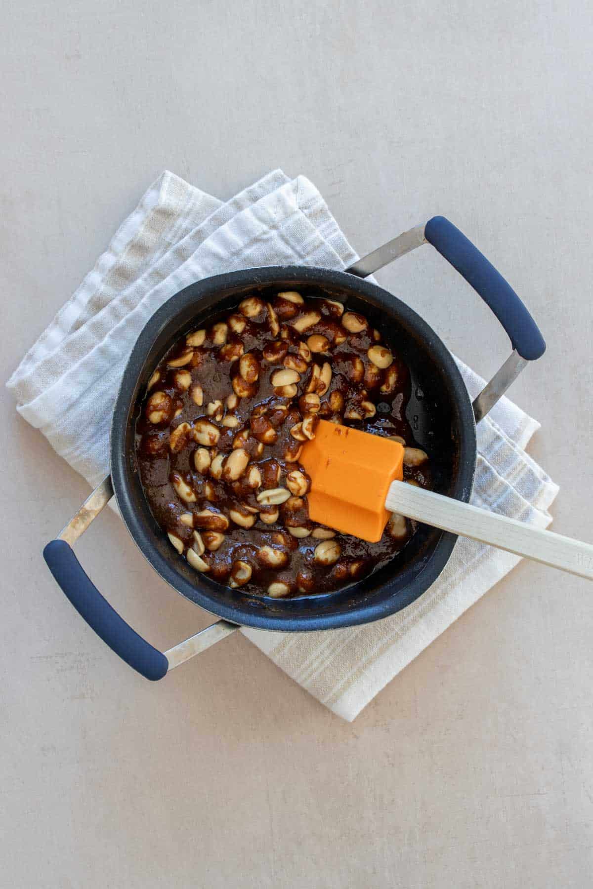 A caramel like mixture with peanuts in a pot being mixed with an orange spatula