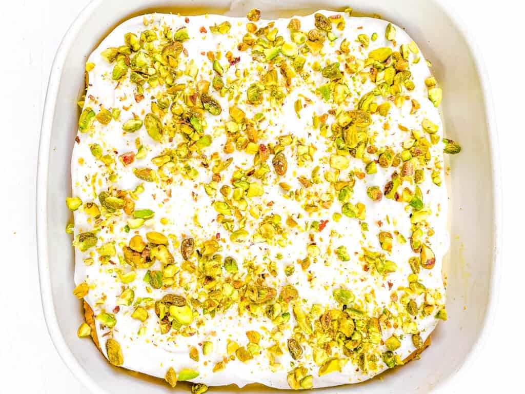 Vegan ras malai tres leches cake in a baking dish, topped with coconut whipped cream and pistachios.