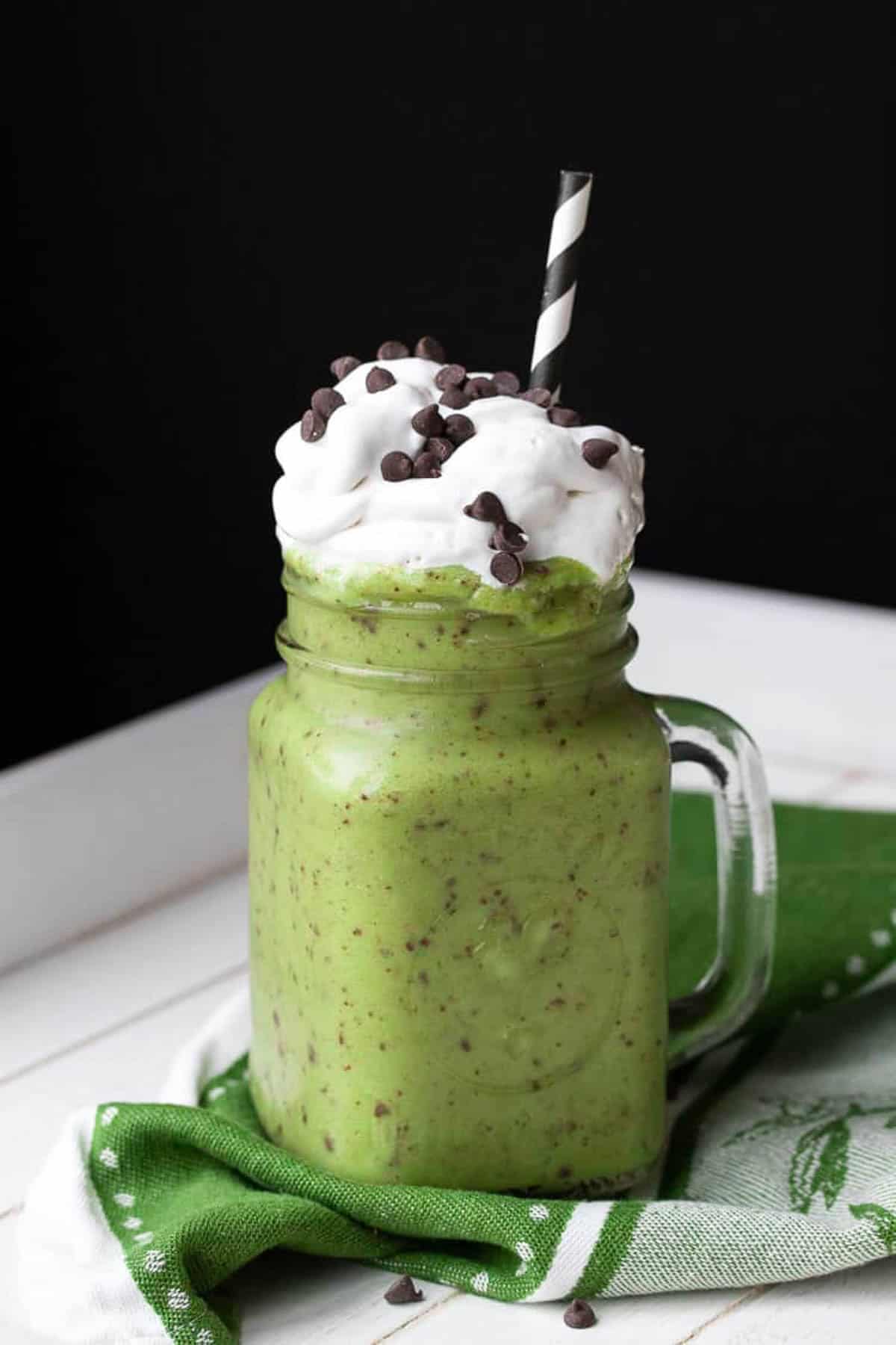 Vegan shamrock shake with chocolate chips in glass jar topped with whipped cream and a straw.